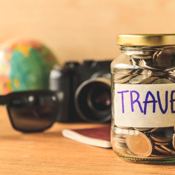 glass jar full of coins with sunglasses, globe, and camera as background