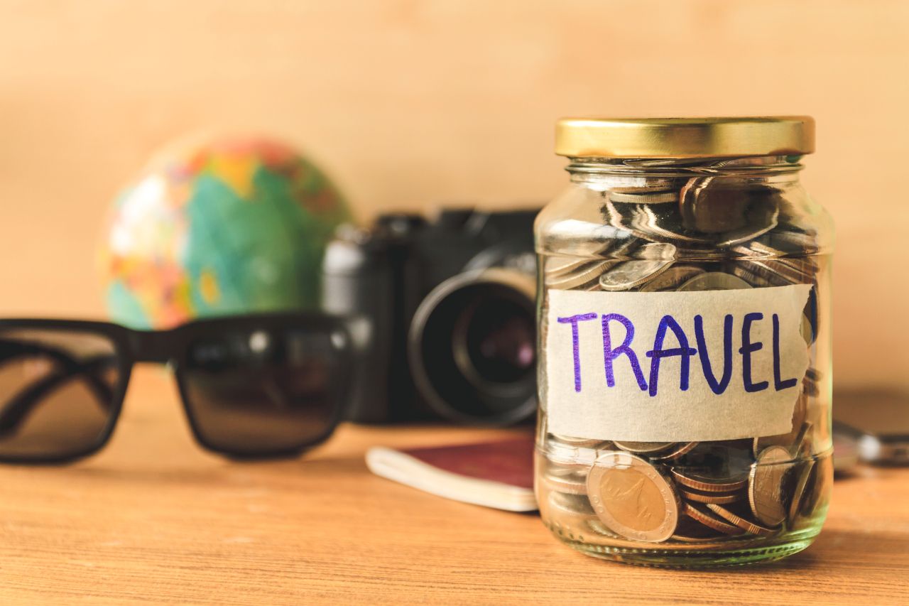 glass jar full of coins with sunglasses, globe, and camera as background