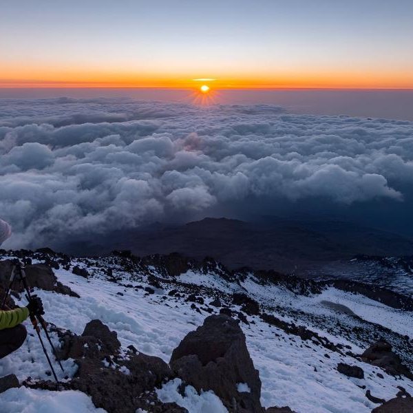 Hiker pauses on snowy mountain peak watching sunrise over clouds
