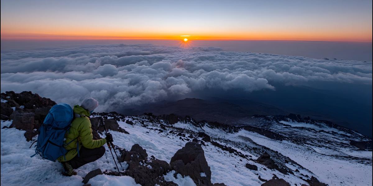 Hiker pauses on snowy mountain peak watching sunrise over clouds